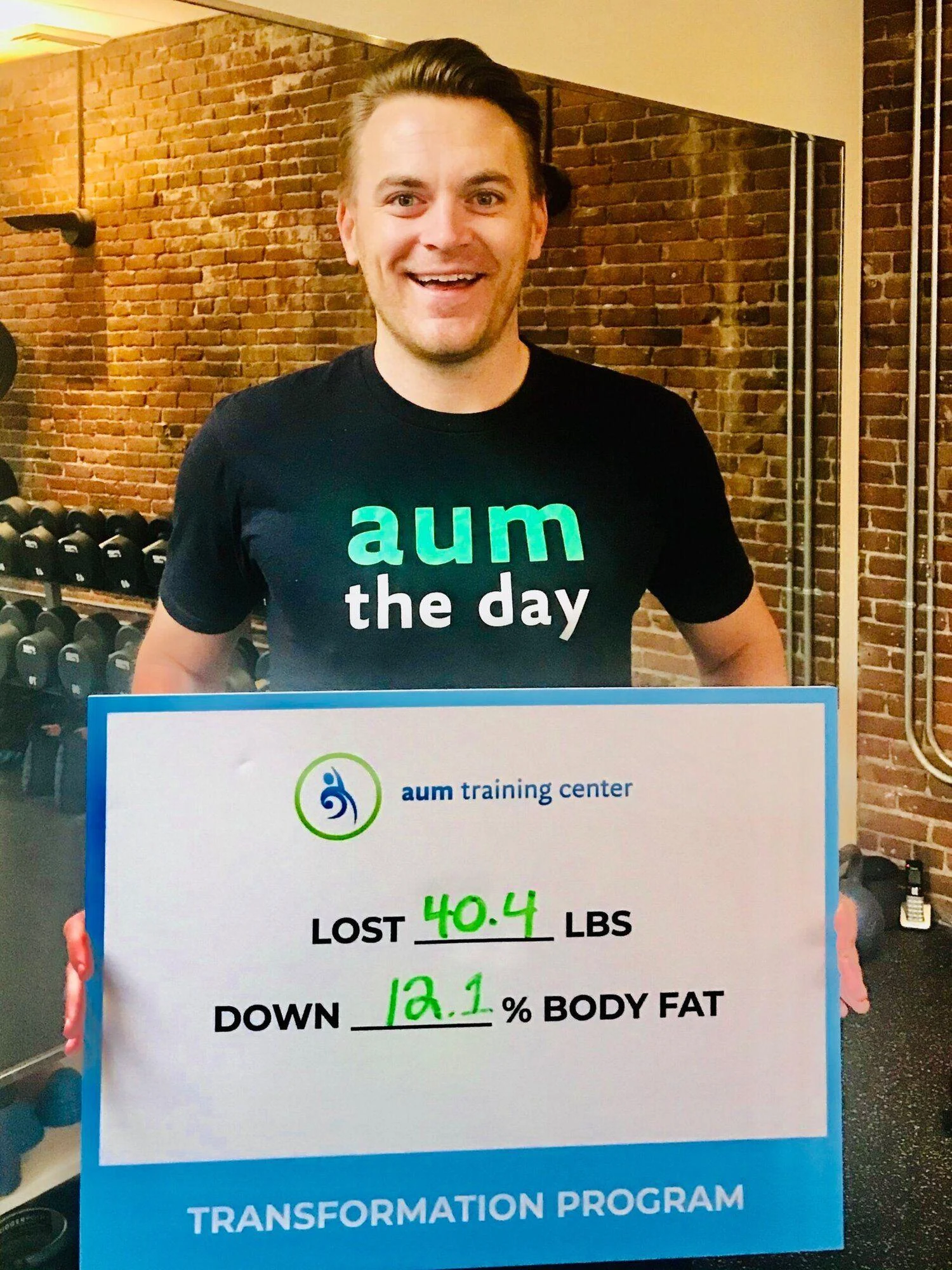 A man stands in a gym holding a sign that reads, "Lost 40.4 lbs, down 12.1% body fat," as part of an aum training center transformation program.