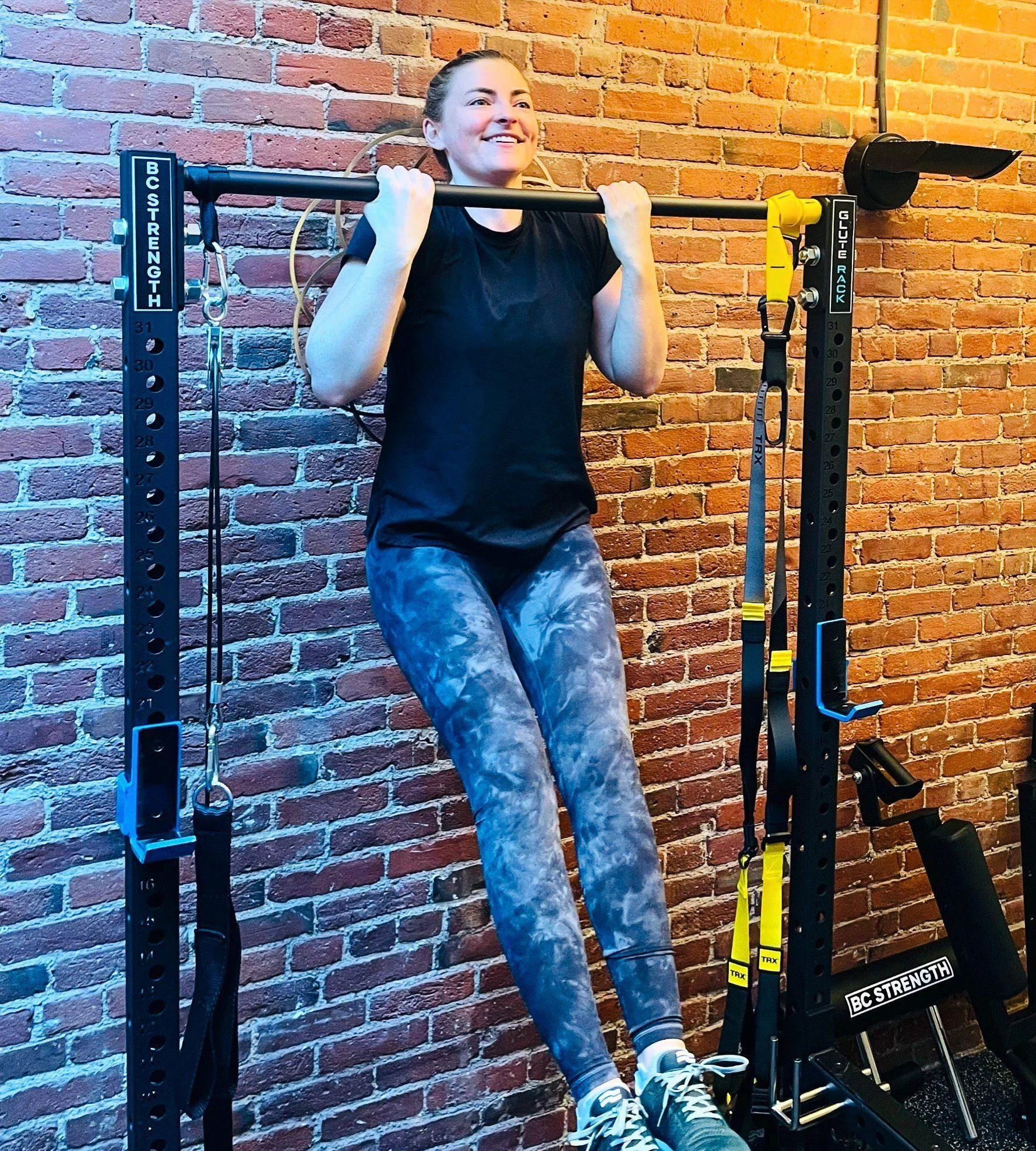 A woman performs a pull-up on a black pull-up bar in a gym with a brick wall background, wearing a black shirt and patterned leggings.