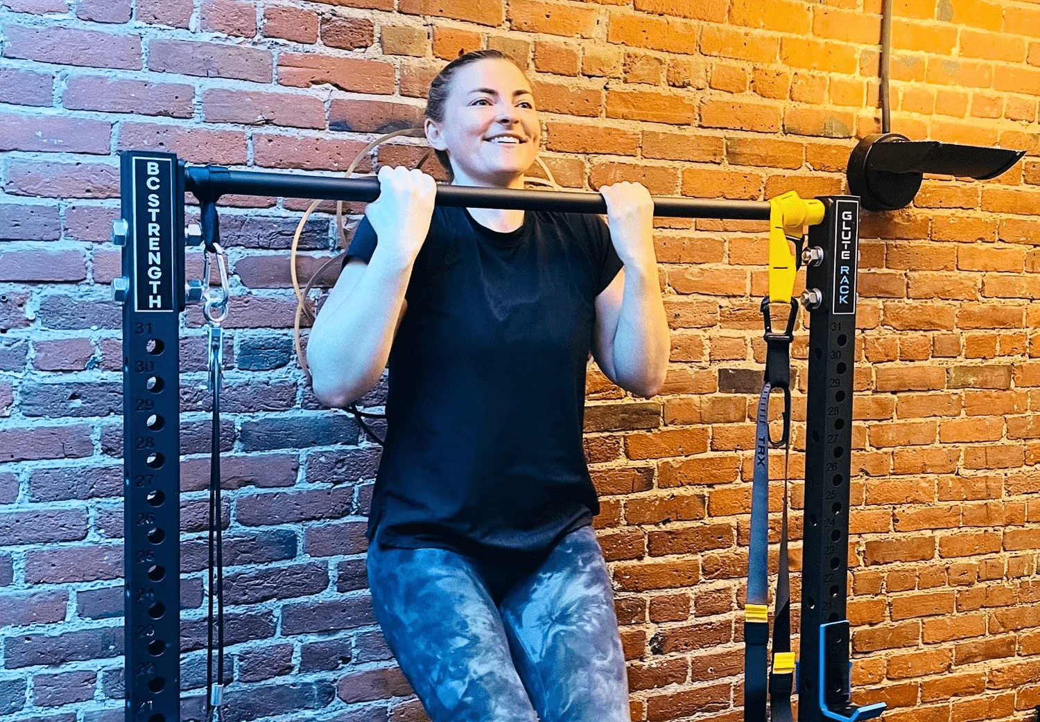 A person wearing a black shirt and patterned leggings performs a pull-up on a BC Strength pull-up bar against a brick wall.
