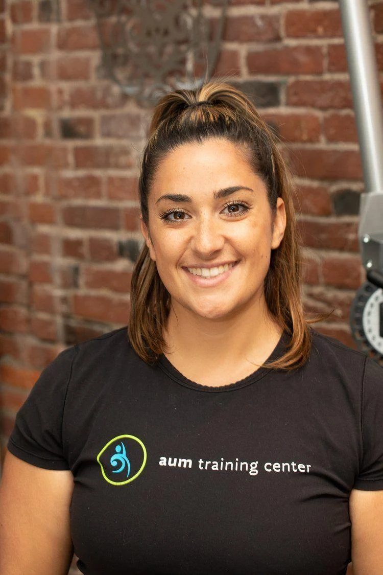 A woman with brown hair in a ponytail, wearing a black "aum training center" t-shirt, smiles while standing in front of a brick wall.