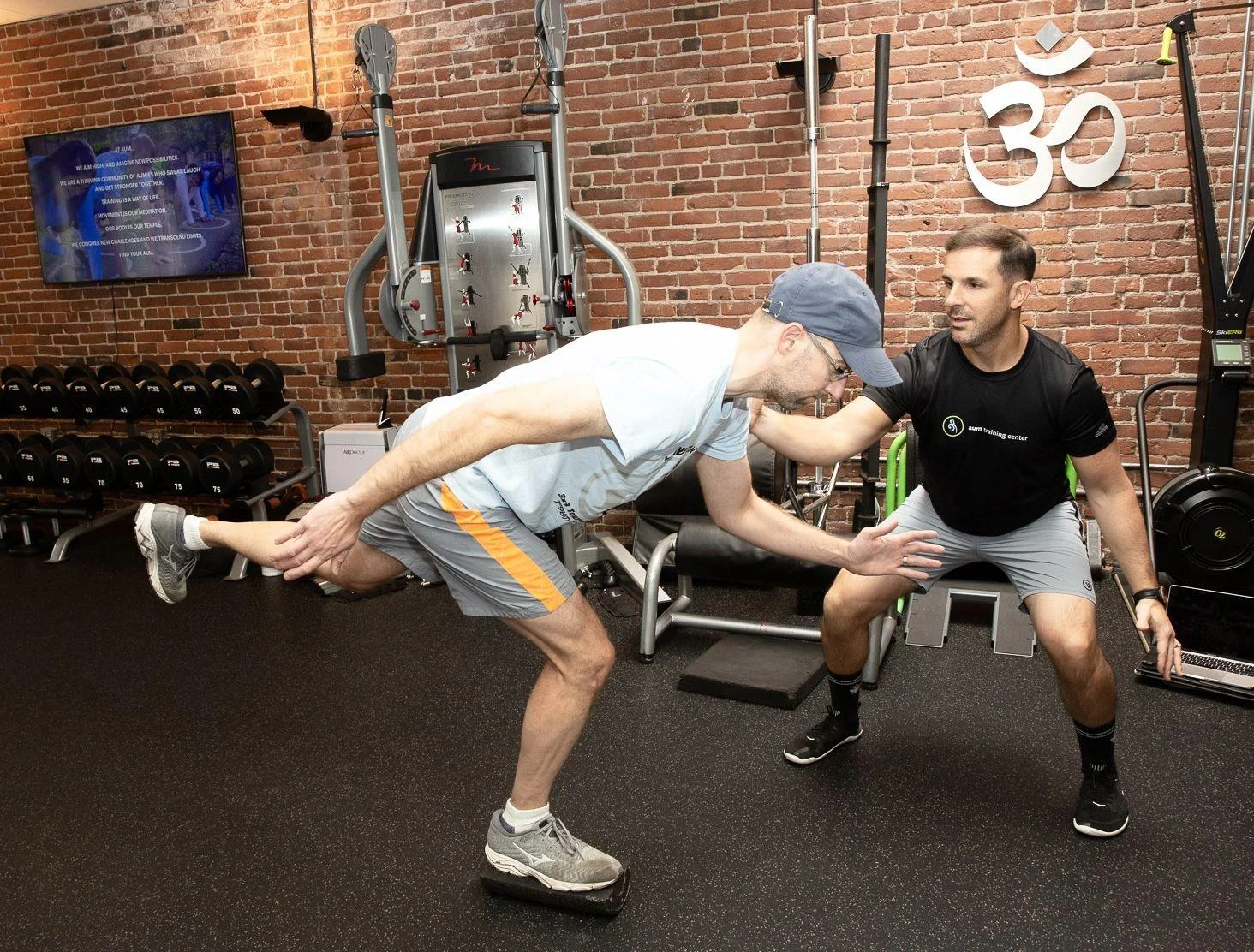 A man balances on one leg atop a balance pad while a trainer observes and supports him in a gym with brick walls and fitness equipment.