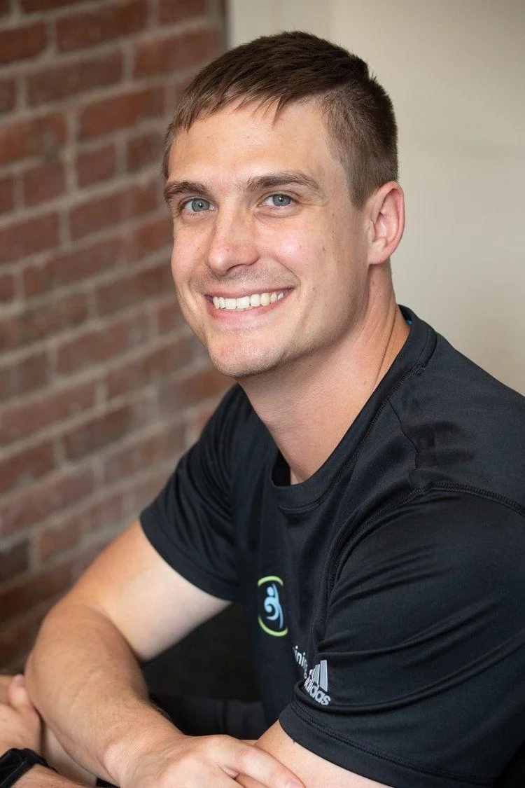 A man with short brown hair wearing a black Adidas shirt smiles while sitting indoors in front of a brick wall.