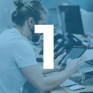 A person with a beard and a man bun sits at a desk using a tablet, with computer monitors in the background and a large number one overlaid on the image.