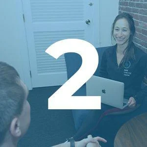 A woman sits with a laptop on her lap, smiling at a person sitting across from her. The number 2 is overlaid on the image.