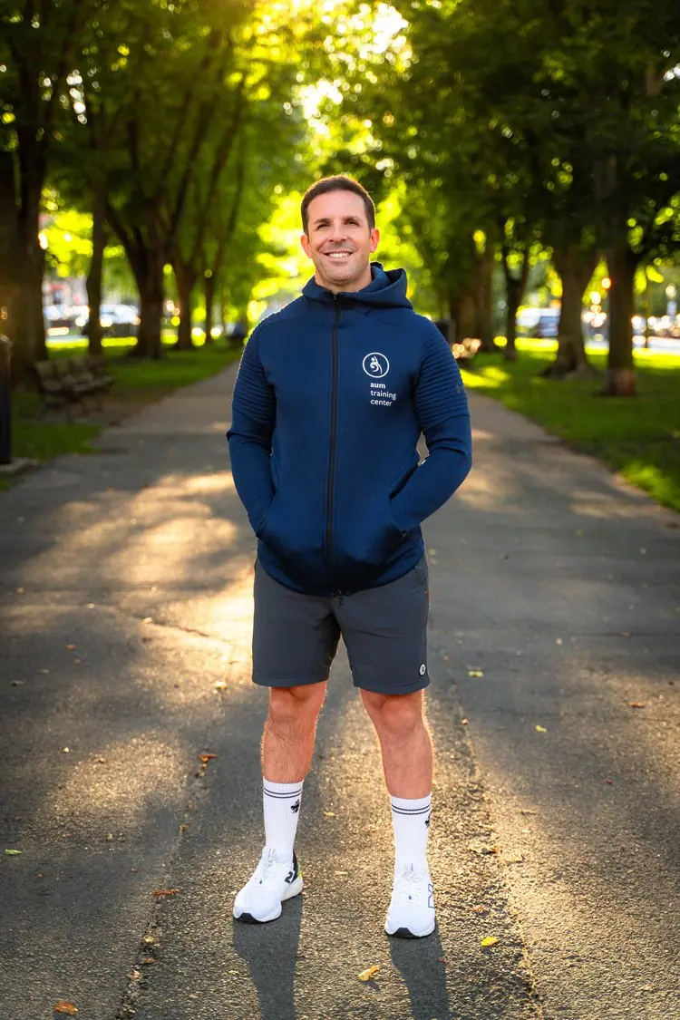 Chris+Carreiro+Headshot+1+low+res Man standing on a sunlit park path, wearing a blue hoodie, gray shorts, and white socks and sneakers, with hands in hoodie pockets and trees in the background.