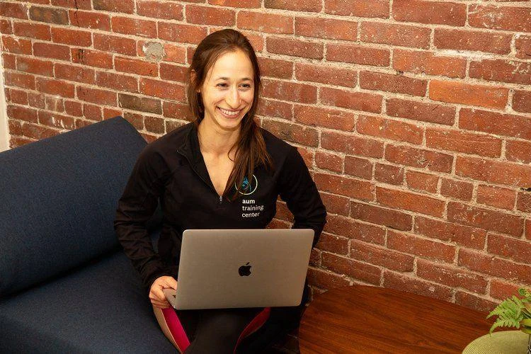 DD774D6C-0CE2-4C8C-A987-4FA1AE3D623A A woman sits on a blue couch with a laptop on her lap, smiling, in front of a brick wall and beside a wooden table with a potted plant.