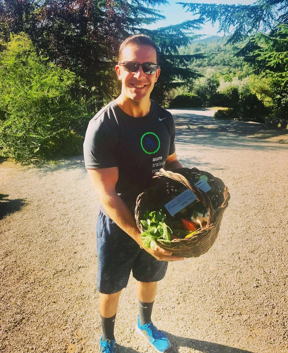 A man wearing sunglasses and athletic clothing stands outdoors holding a wicker basket filled with fresh vegetables.
