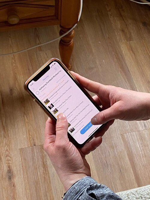 IMG_3539 A person holds a smartphone displaying a list of messages or notifications on the screen while standing on a wooden floor near a piece of furniture.