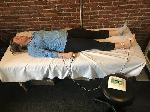 IMG_4469 A person lies on a table with electrodes attached to their hands and feet, connected to a small electronic device displaying numbers.