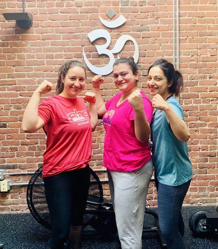 IMG_7708+2 Three women stand side by side in front of a brick wall with an Om symbol, flexing their arms and smiling, wearing athletic clothing.