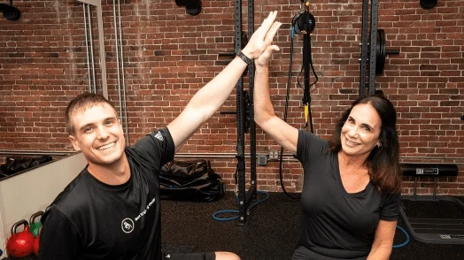 Image 21 A man and a woman in athletic wear high-five each other in a gym setting with brick walls and exercise equipment in the background.