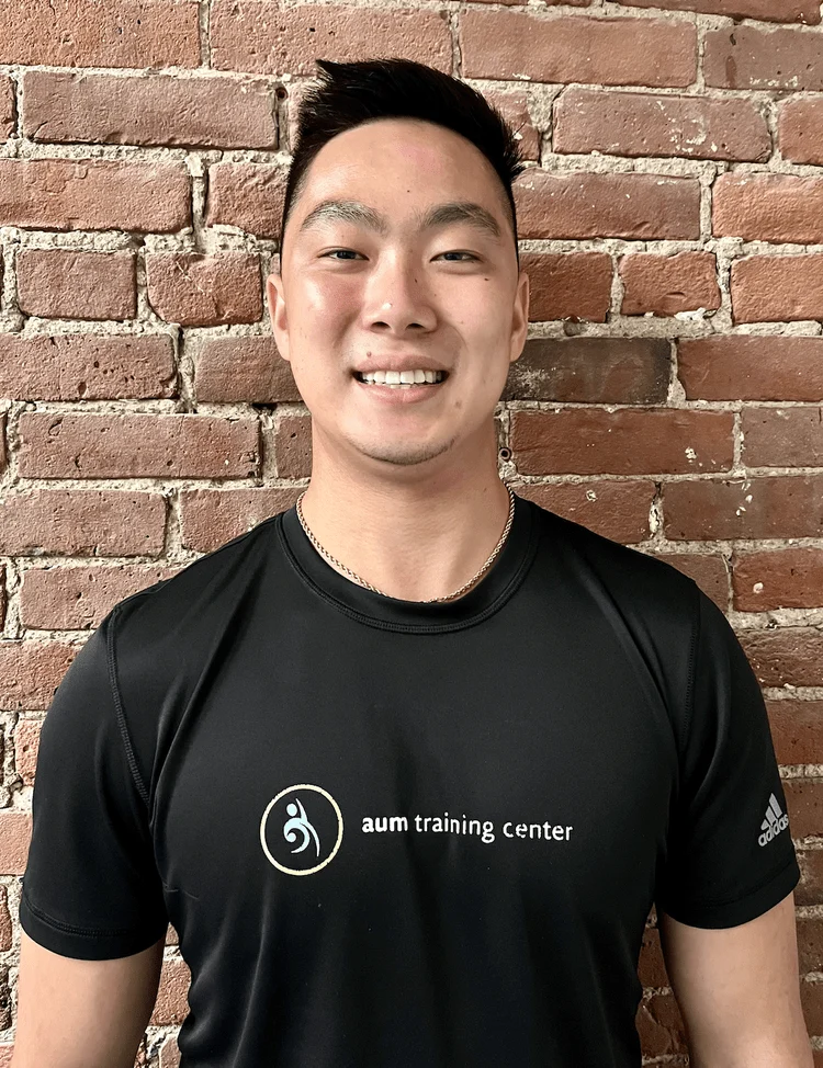 Screen+Shot+2024-07-16+at+3.50.24+PM A person stands smiling in front of a brick wall, wearing a black "aum training center" t-shirt.