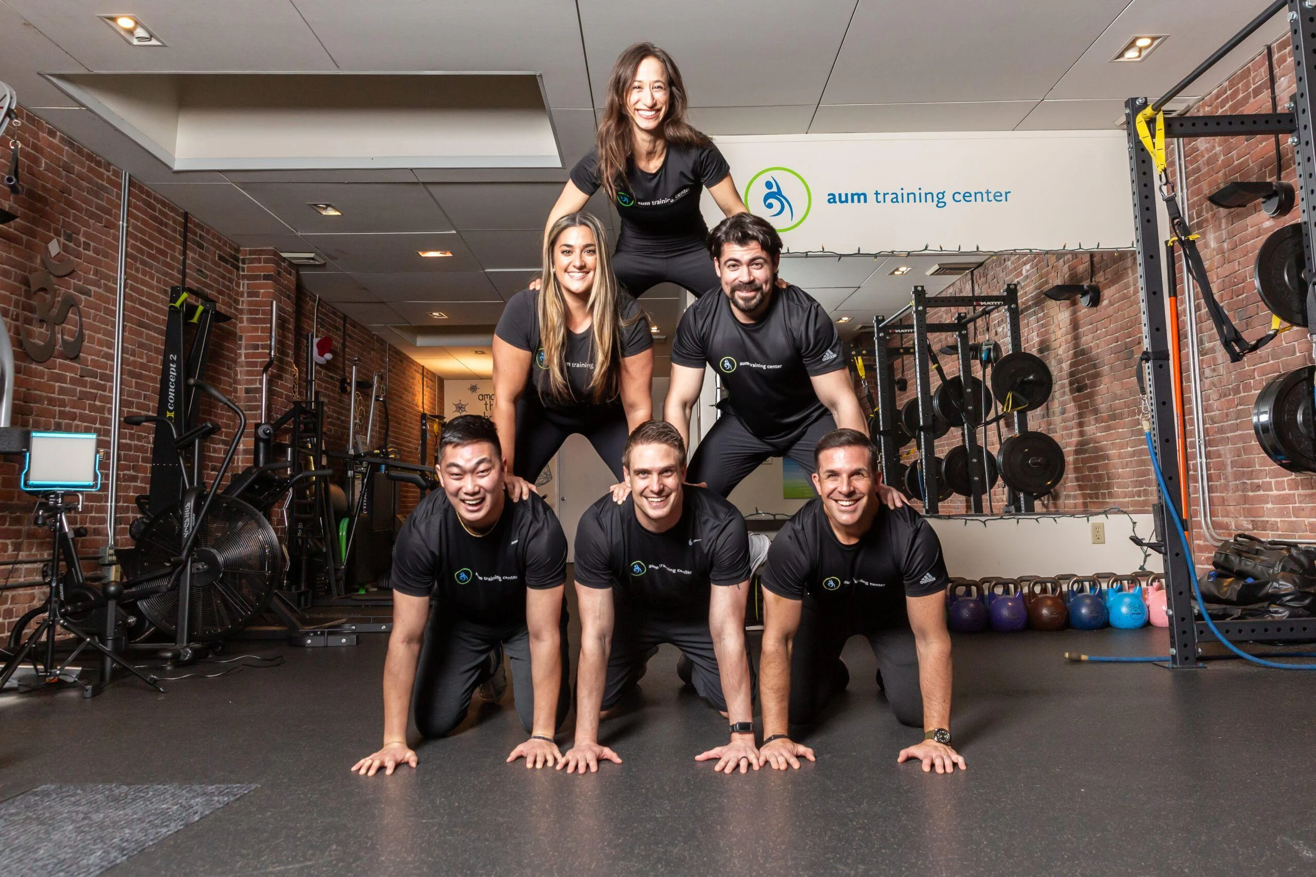 Six people in matching workout attire form a human pyramid inside a gym with brick walls and exercise equipment visible in the background.
