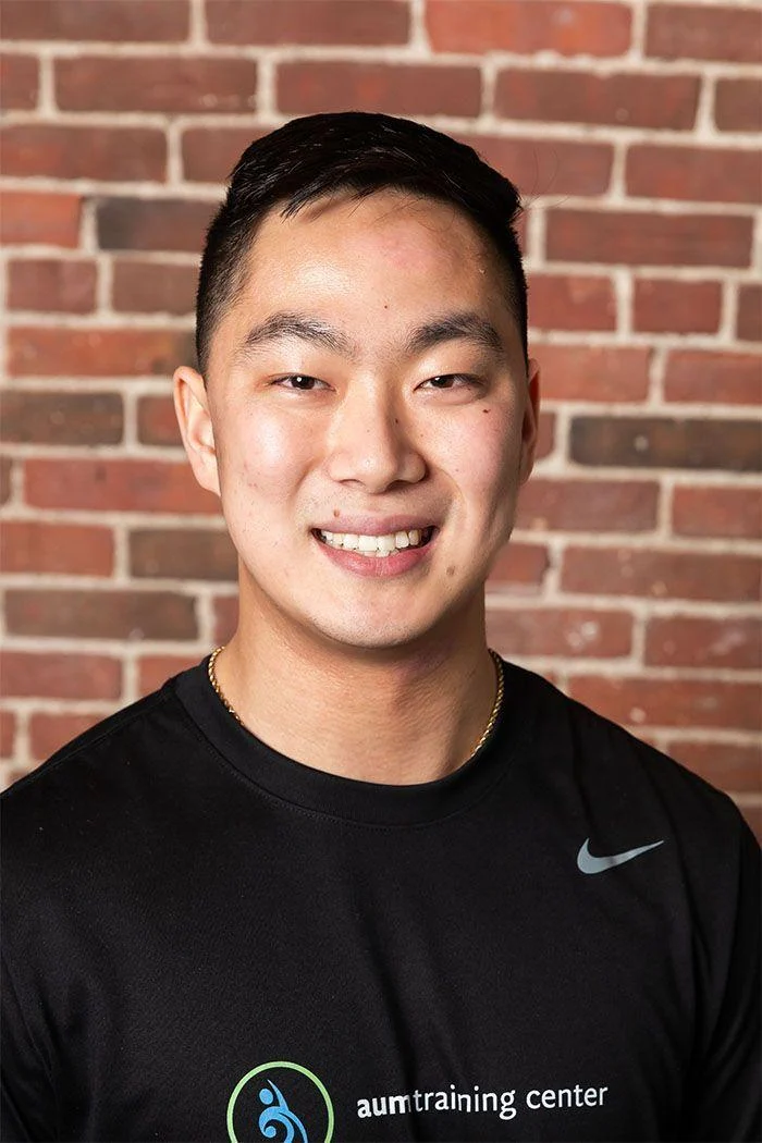 A young man with short black hair smiles at the camera, wearing a black Nike shirt with "aum training center" logo, standing in front of a brick wall.