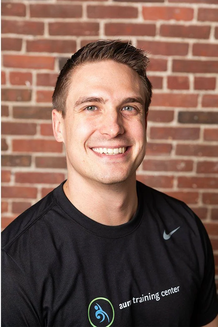Man with short brown hair smiling, wearing a black "aum training center" t-shirt, standing in front of a brick wall.