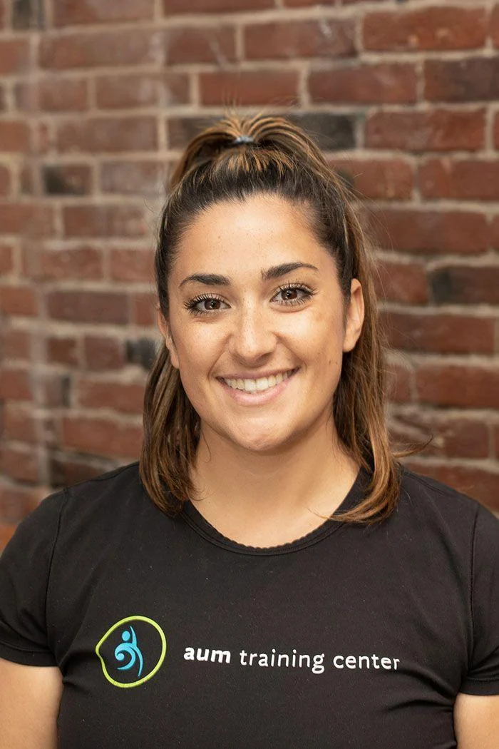 A woman with long brown hair in a ponytail, wearing a black "aum training center" t-shirt, smiles in front of a brick wall.