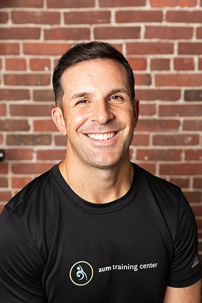 A man wearing a black "aum training center" t-shirt smiles at the camera while standing in front of a brick wall.