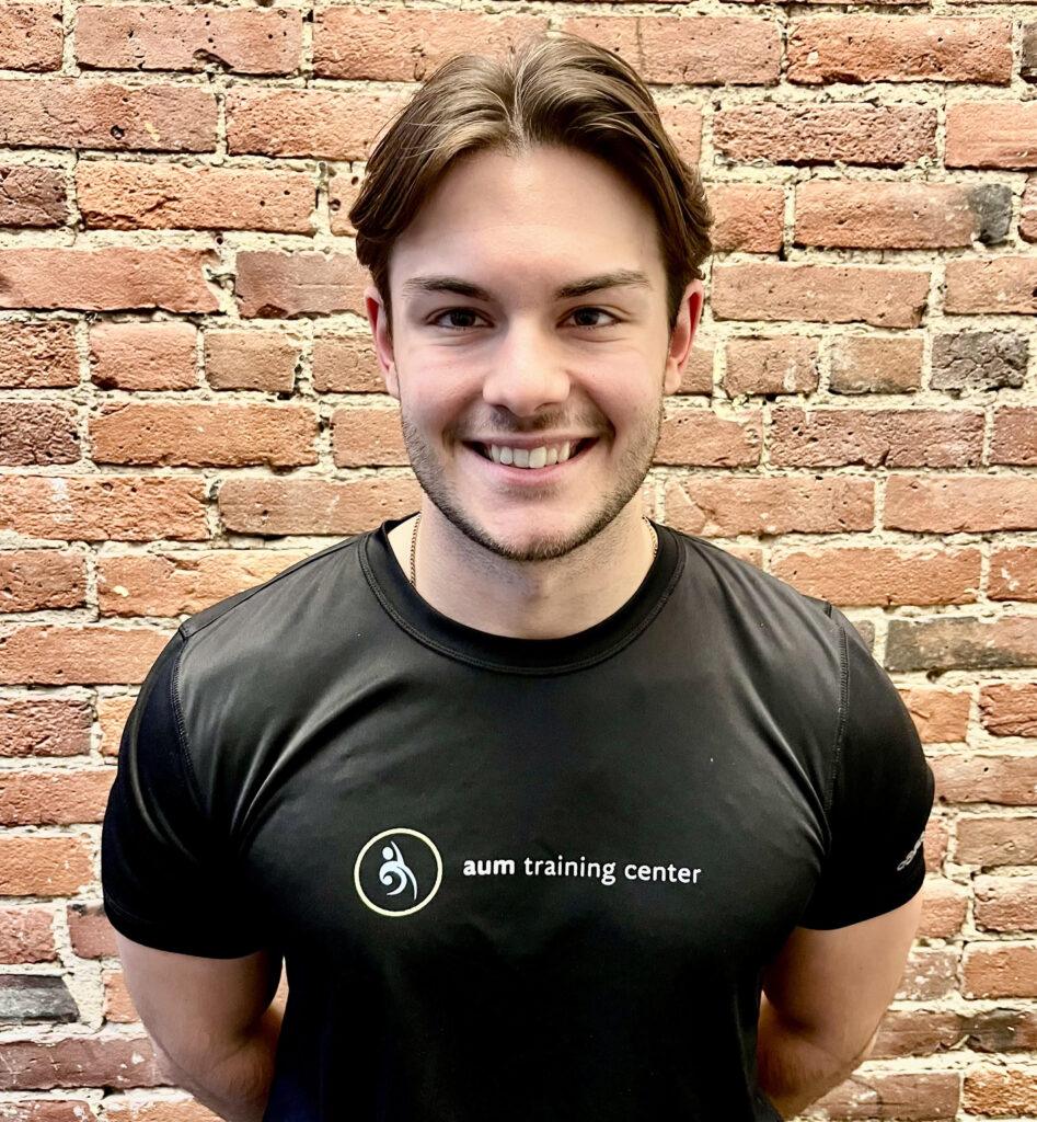 A person smiling and standing in front of a brick wall, wearing a black "aum training center" t-shirt.