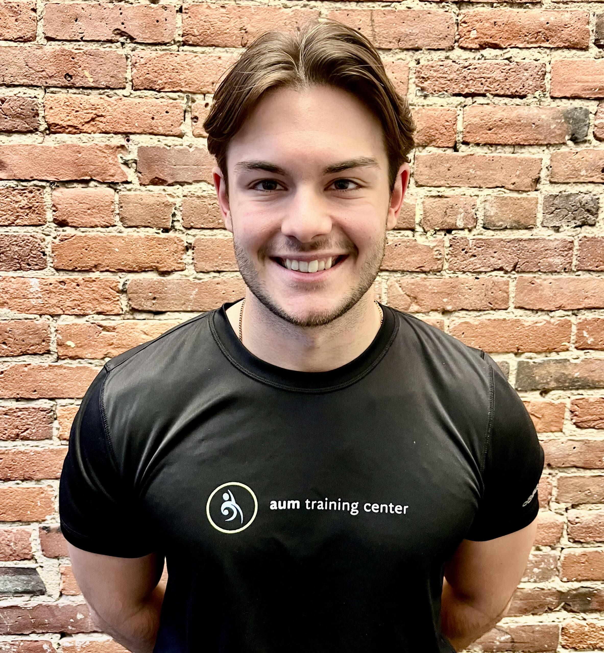 coach-max-aum-fitness A person smiling and standing in front of a brick wall, wearing a black "aum training center" t-shirt.