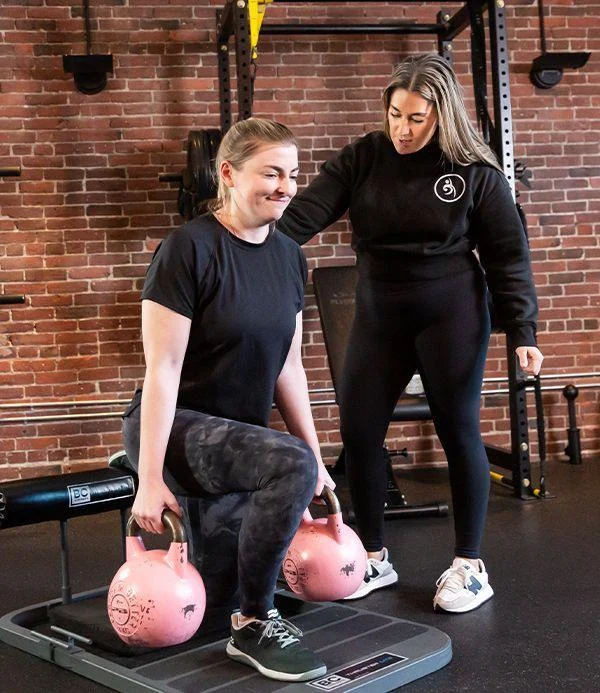 A woman performs a lunge exercise with pink kettlebells while another woman, likely a trainer, observes and provides guidance in a gym with exposed brick walls.