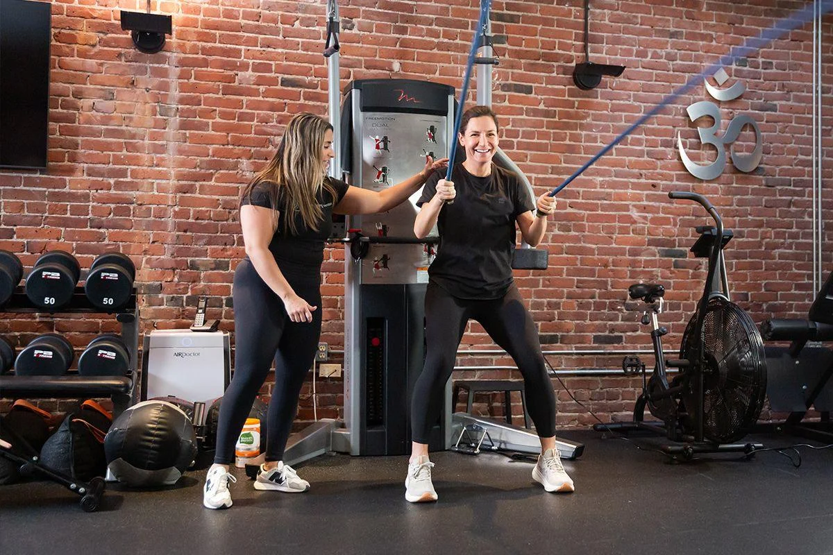 energylast Two women exercise in a gym; one is using resistance bands while the other provides guidance. Gym equipment and weights are visible in the background against a brick wall.