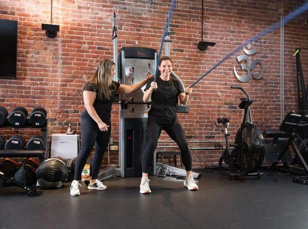 equipped Two women exercise in a gym; one uses resistance bands while the other, possibly a trainer, assists her. Gym equipment and weights are visible against a brick wall background.