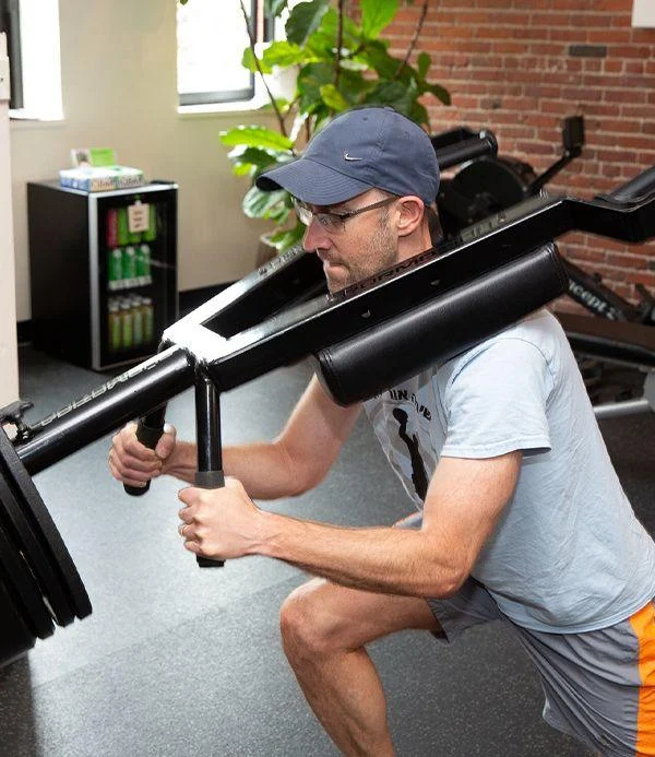 A man wearing a cap and glasses performs a squat using a loaded safety squat bar in a gym setting.