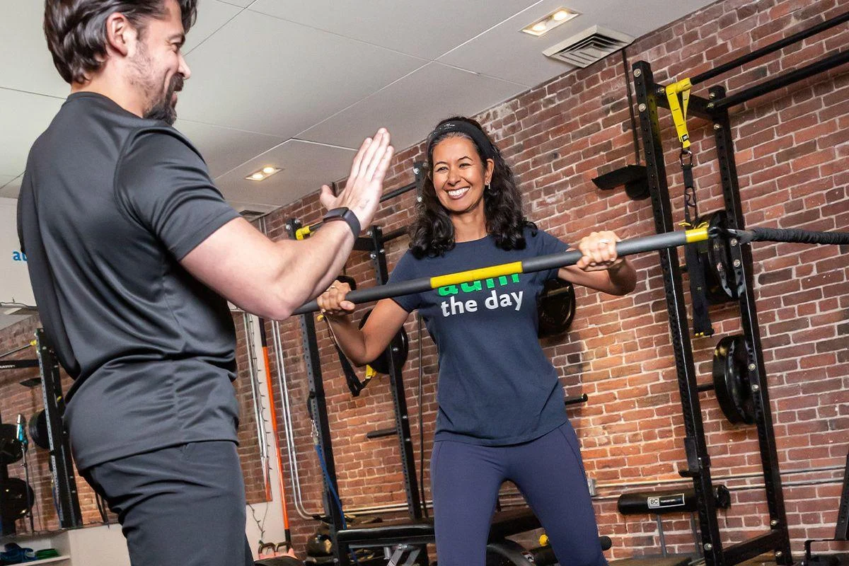 events2 A woman lifting a barbell smiles as she receives a high-five from a man in a gym with brick walls and fitness equipment.