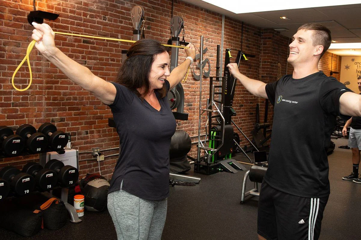 fat3 A woman exercises with a resistance band, supervised by a trainer, in a gym with brick walls and various fitness equipment in the background.