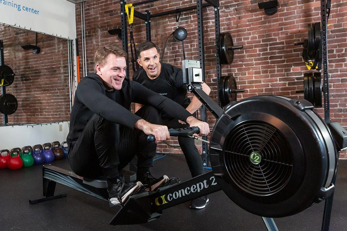 fat34 Two men are in a gym; one is using a Concept2 rowing machine while the other stands behind, observing and offering guidance.