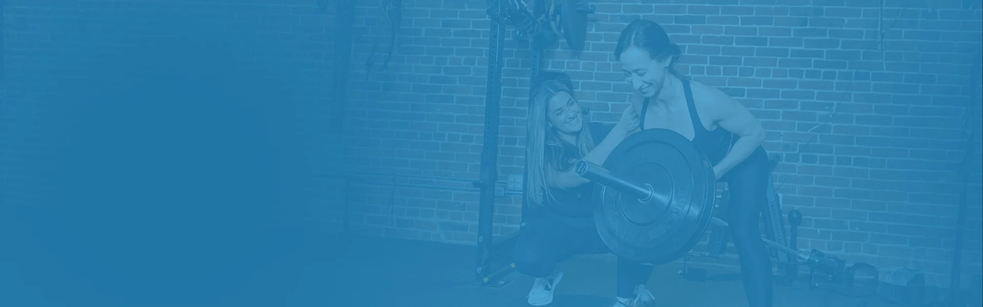 A woman in athletic wear uses a gym cable machine while a trainer stands behind her, providing guidance and support. Fitness equipment is visible in the background.