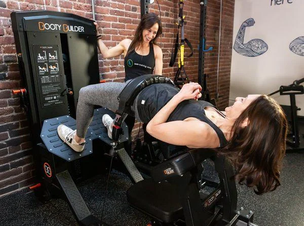 feature-2 Two women are in a gym; one is using a leg press machine labeled "Booty Builder" while the other stands nearby, smiling and assisting.