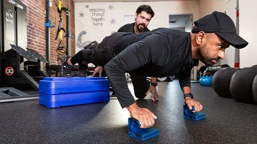 habits Two men exercising in a gym; one man does push-ups using handles while the other crouches nearby, observing and coaching. Gym equipment is visible in the background.
