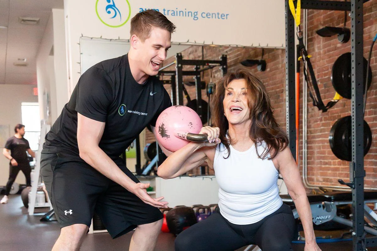 health-2 A woman in athletic wear does a kettlebell squat while a male trainer encourages her at a gym with various equipment in the background.