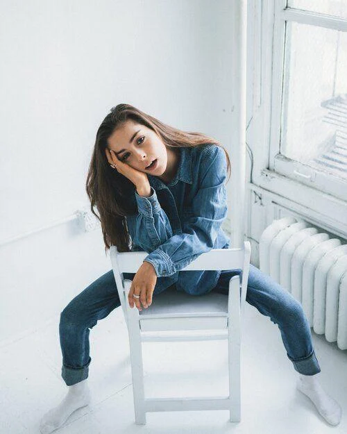 image-asset A woman wearing denim jeans and a denim shirt sits backward on a white chair in a bright room, resting her head on one hand and looking at the camera.