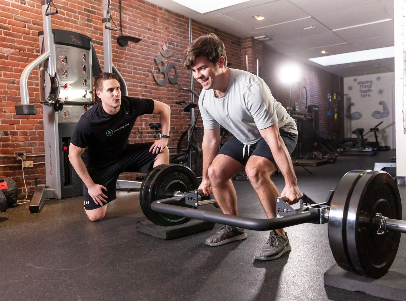 personal-training-1 A man deadlifts a barbell while another man kneels nearby, observing him in a gym with brick walls and various exercise equipment.