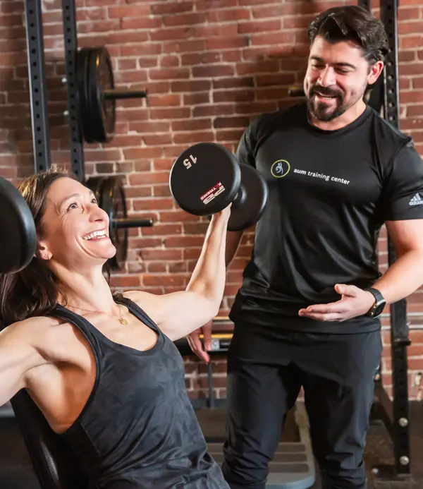 personal-training-1 A woman lifts a dumbbell while seated on a bench, smiling, as a trainer stands beside her in a gym with brick walls and black equipment.