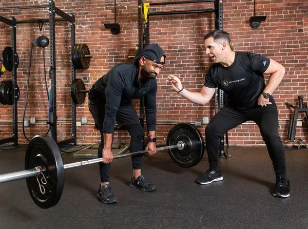 personal-training Two men are in a gym; one is preparing to lift a barbell while the other, likely a trainer, gestures and encourages him.