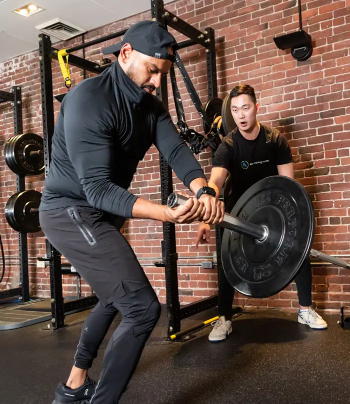 personal A man performs a barbell exercise in a gym while another man stands behind him, observing and providing support.