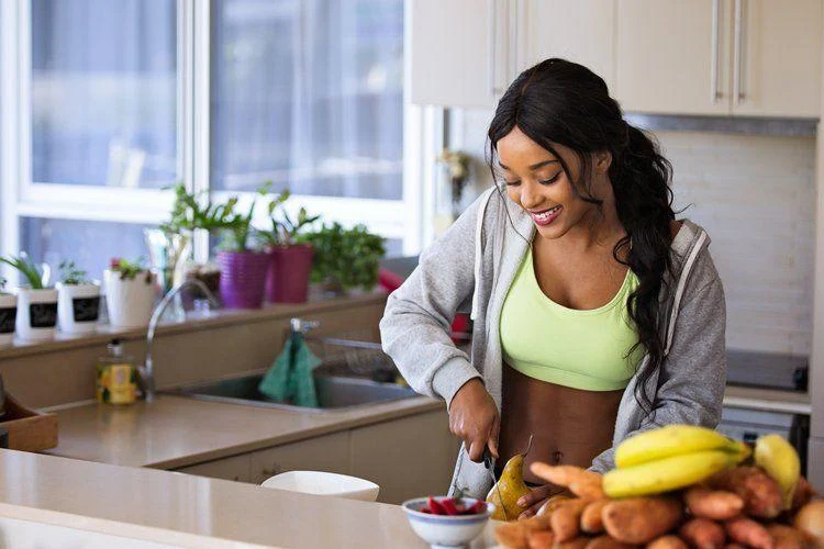 pexels-nathan-cowley-1153369 A woman in athletic wear smiles while slicing food in a bright kitchen. Bananas and sweet potatoes are on the counter, and potted plants are in the background.