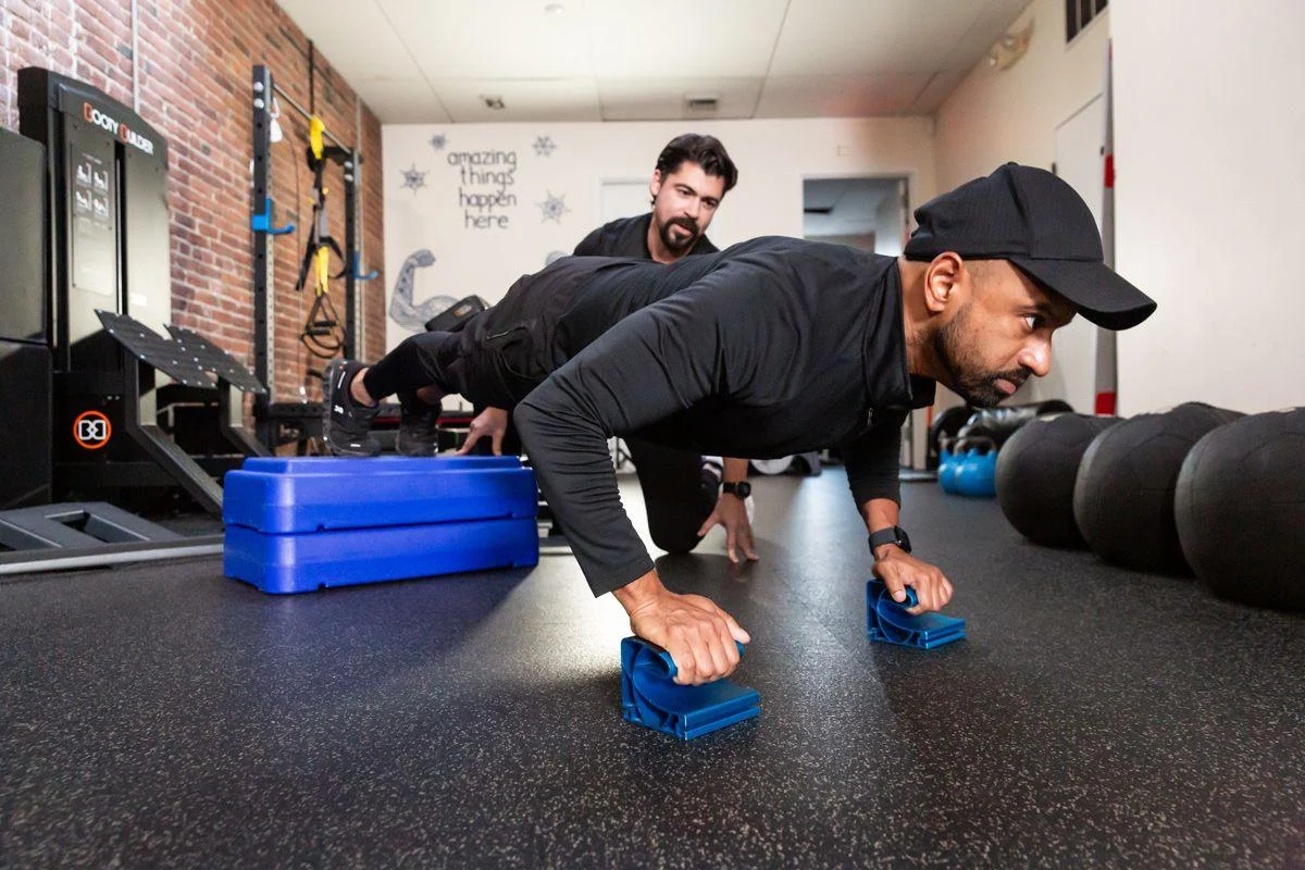 strength A man performs push-ups using push-up bars in a gym while a trainer observes. Exercise equipment and kettlebells are visible in the background.