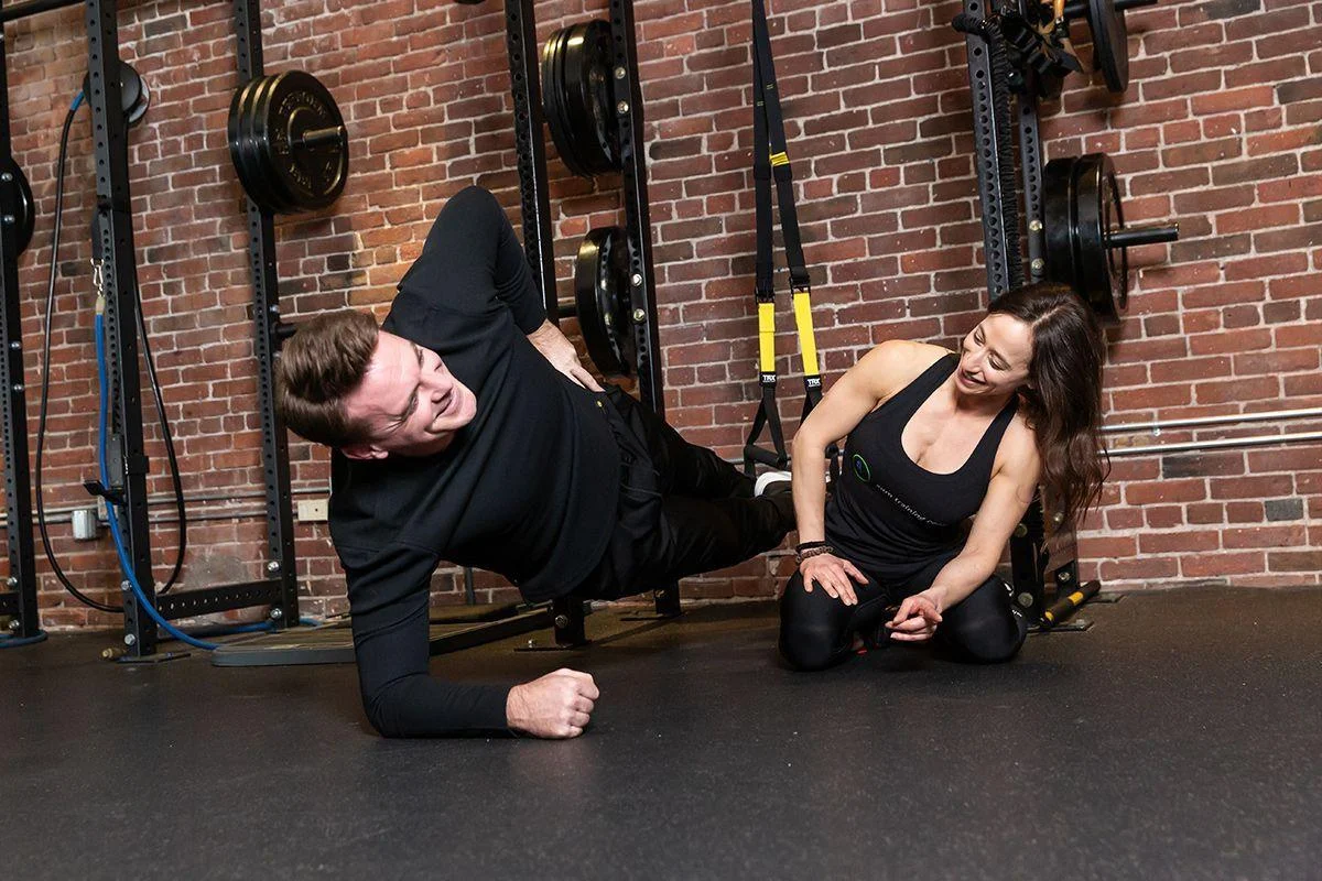 t32 A man performs a side plank exercise on the gym floor while a woman, possibly a trainer, kneels nearby and watches him. Gym equipment and a brick wall are in the background.