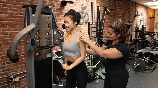 t3333 A woman assists another woman in using a cable machine at a gym with brick walls and various exercise equipment in the background.
