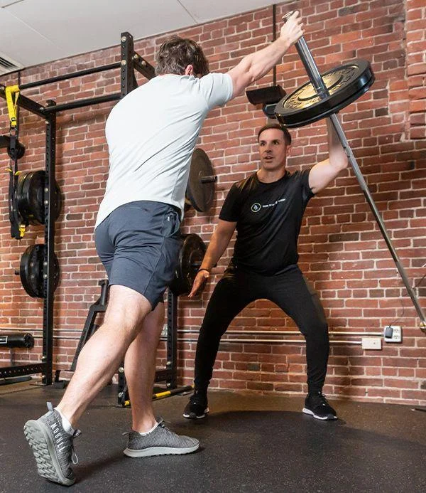 transf1 Two men exercise in a gym; one pushes a weighted barbell anchored at the floor while the other supervises. Brick wall and gym equipment are visible in the background.