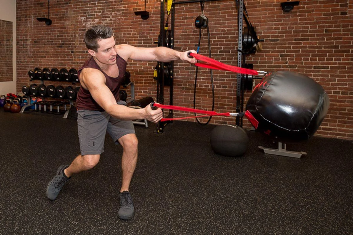 transform A man in athletic wear exercises in a gym, pulling resistance bands attached to a large swinging bag.