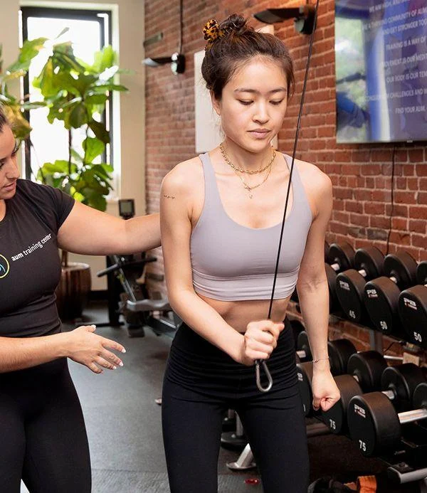 A woman performs a cable exercise in a gym while a trainer assists her. Dumbbells are visible in the background.