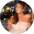 unnamed Woman with long brown hair wearing a strapless beige top sits indoors, holding an orange drink, with city lights visible through the window behind her.