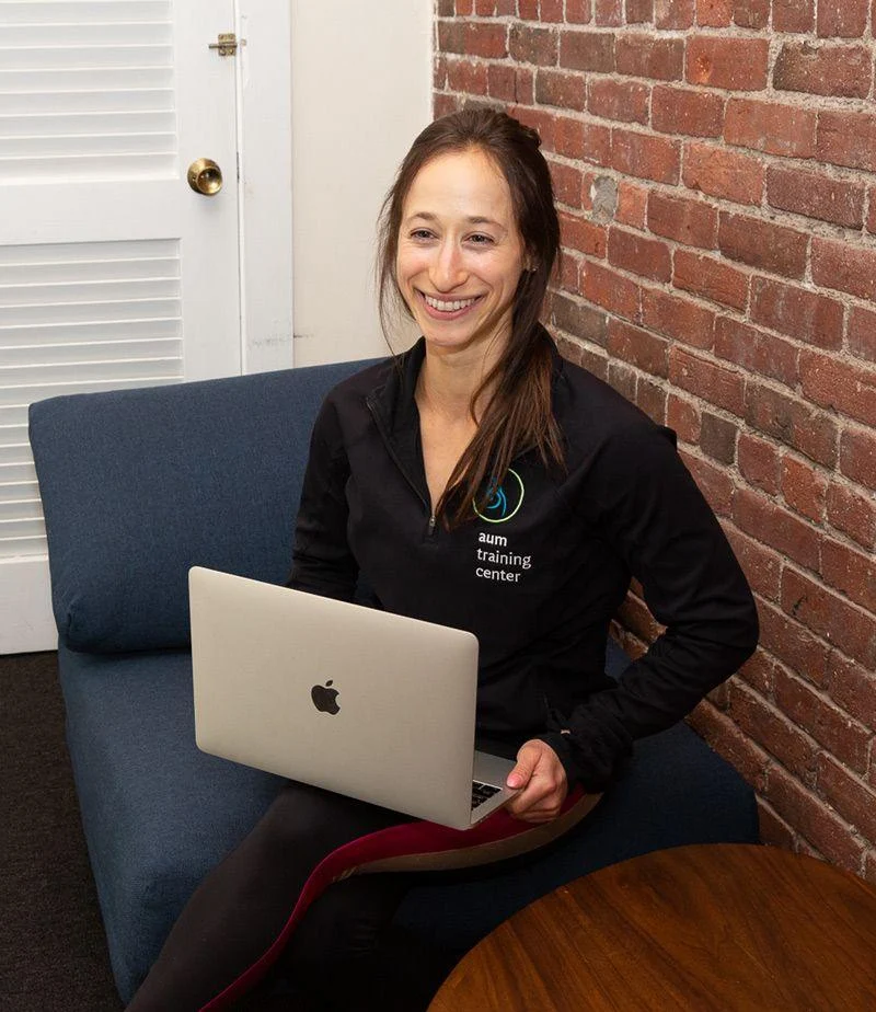 virtual-2 A woman sits on a blue couch with a laptop on her lap, smiling. She wears a black jacket with "training center" text on it. A brick wall and white door are in the background.