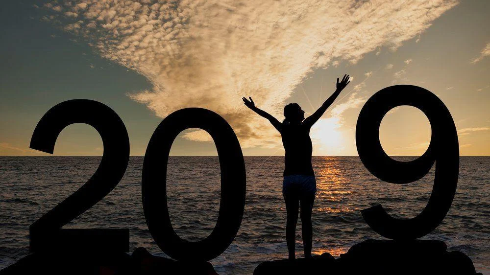 GettyImages-1038621346 A person stands with arms raised between large “2019” numbers at the beach during sunset, with clouds and the ocean in the background.