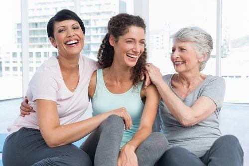 Cheerful fit women in yoga class Three women of different ages sit close together, smiling and laughing in a bright indoor setting.